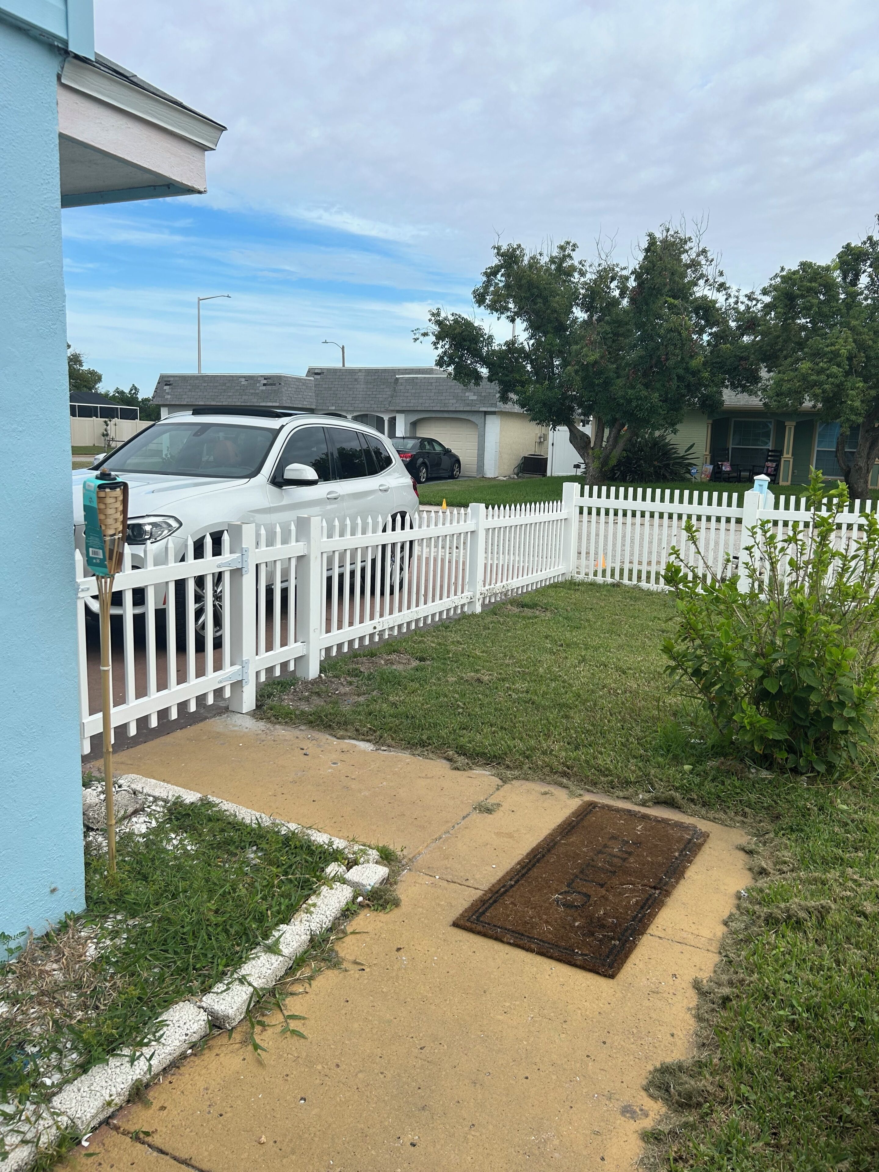 Un barrio residencial con una cerca blanca enrejada, un automóvil blanco estacionado al frente y un jardín cubierto de césped.