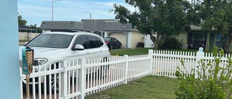 Un barrio residencial con una cerca blanca enrejada, un automóvil blanco estacionado al frente y un jardín cubierto de césped.