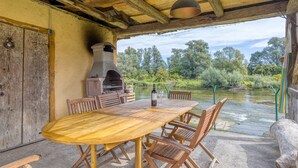 An outdoor patio with a wooden table and chairs, a stone oven, and a view of a river and trees.