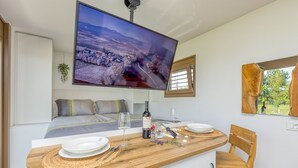 A modern room with a bed, a TV mounted on the wall, a wooden dining table with chairs, and a window with a view of greenery.