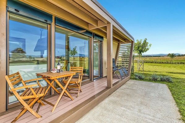 A modern outdoor patio with a wooden table and chairs, large sliding glass doors, and a view of a green landscape.