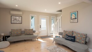 Ein Wohnzimmer mit zwei grauen Sofas, einem Couchtisch und einem Teppich. Zwei Türen führen in andere Räume, und ein gerahmtes Bild hängt an der Wand.