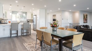 Una cocina moderna con gabinetes blancos, un refrigerador de acero inoxidable y un comedor con mesa y sillas de vidrio.