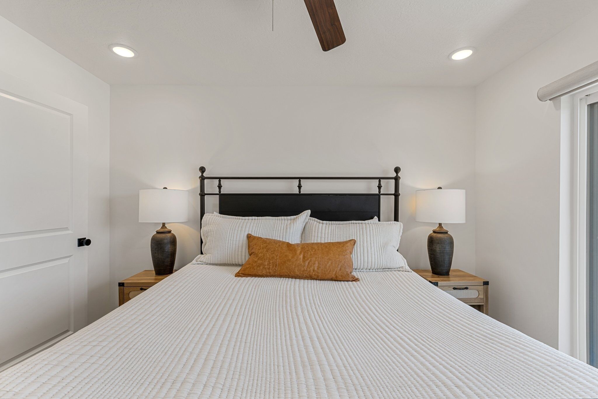 A bedroom with a large bed, two bedside tables with lamps, a ceiling fan, and a window with blinds.