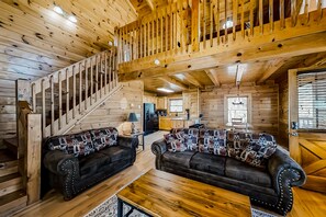 Cozy cabin interior with a high ceiling and exposed beams, featuring a living area with two sofas and a kitchen area with wooden cabinets.