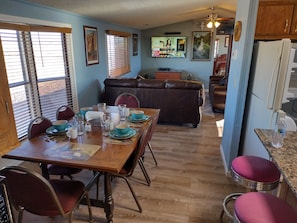 Un comedor con una mesa de madera preparada para cuatro personas, rodeada de sillas. Un sofá de cuero marrón se encuentra en el fondo, y hay un televisor montado en la pared.