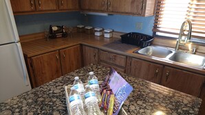 A kitchen with granite countertops, wooden cabinets, a stainless steel sink, and a basket of bottled water and snacks.