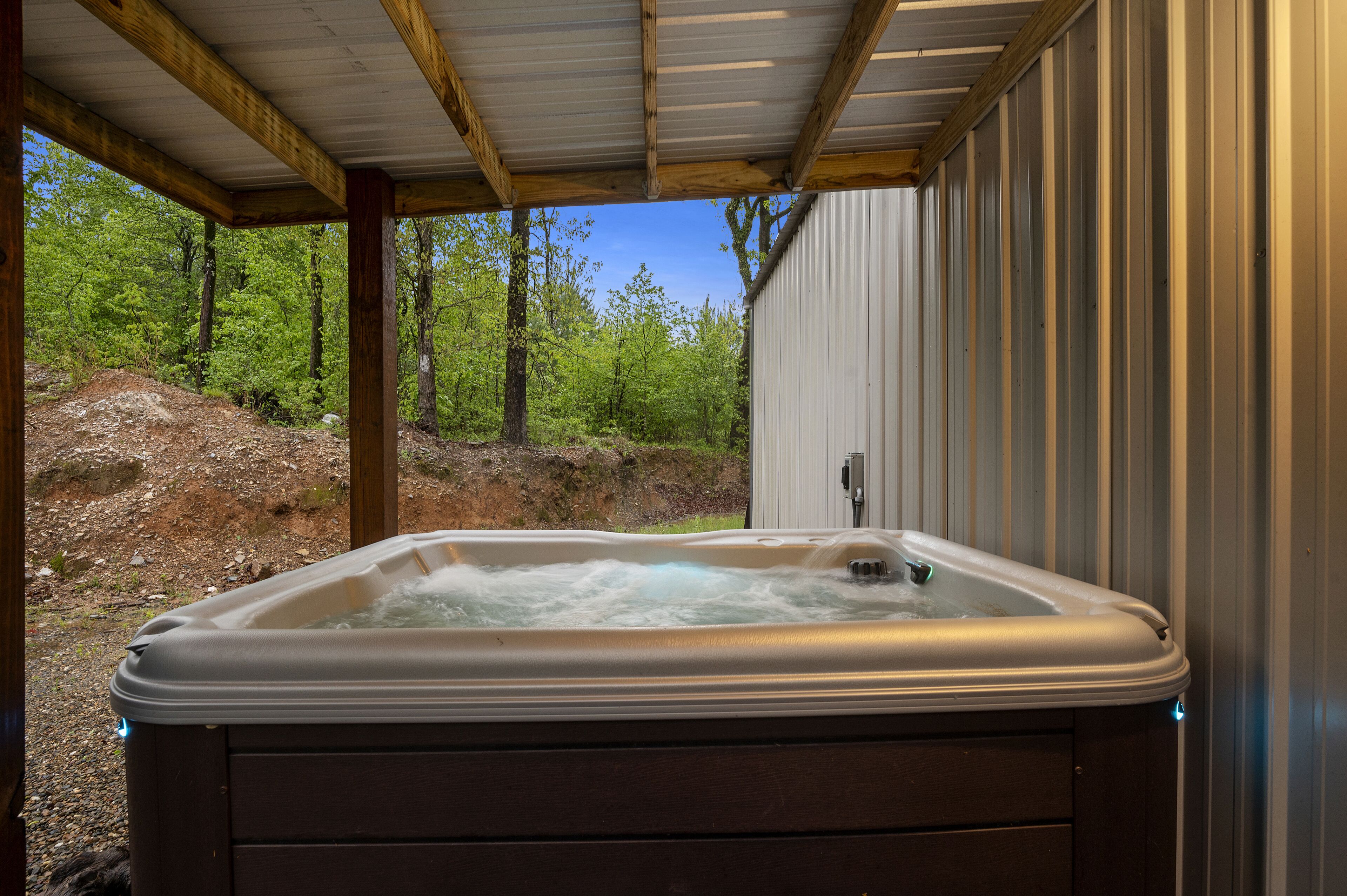 Een hottub in een open omgeving, met een houten terras en metalen wanden, omgeven door bomen en een rotsachtig landschap.