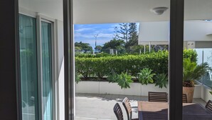 A view through a glass door to an outdoor patio area with a table and chairs, surrounded by greenery and a clear sky.