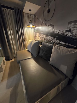 Un dormitorio moderno con un mural de paisaje urbano, una cama con almohadas, una lámpara colgante y una pequeña mesita de madera.