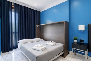 Habitación de hotel moderna con una cama grande, cortinas oscuras, una pared azul de acento, una mesita de noche con una lámpara y una planta pequeña.