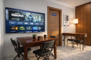 Habitación de hotel con un televisor de pantalla plana grande mostrando una película, una puerta de madera con la leyenda 'Blue room', una mesita con sillas y una lámpara.