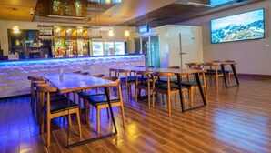 Zona de bar con mesas y sillas de madera, una barra con diferentes botellas y una pantalla de televisión grande en la pared.