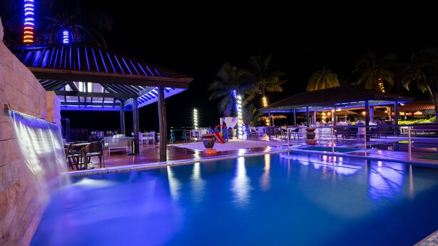 Zona de piscina nocturna con área de descanso cubierta, palmeras y un bar/lounge con gente.