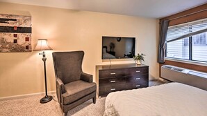 A bedroom with a bed, a dresser, a chair, a lamp, a TV, and a window with blinds.