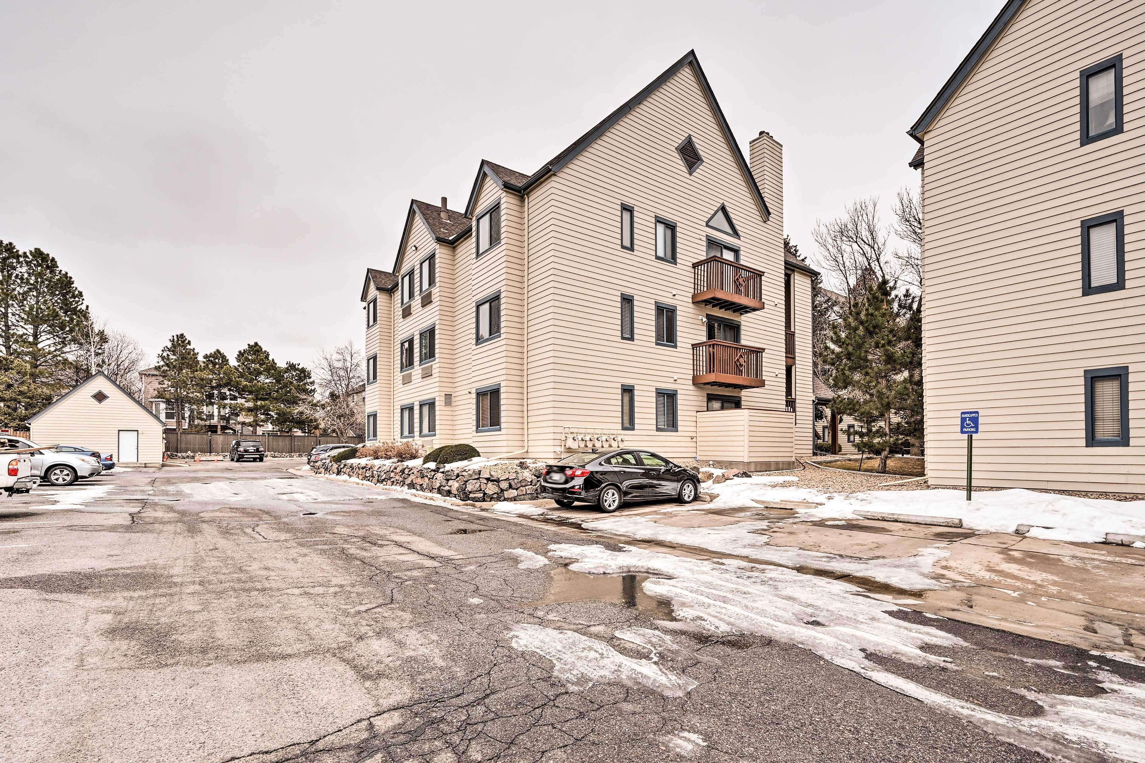A multi-story residential building with a sloped roof, a balcony on the second floor, and a parking area with cars and a snow-covered ground.