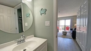A modern bathroom with a white sink, a large mirror, and a door leading to a living area with a sofa and a view of the outside. - Ocean Dunes by Palmetto Vacations (Myrtle Beach)