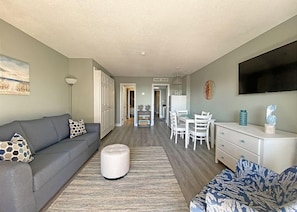 Una sala de estar moderna con un sofá gris, un reposapiés y un sillón estampado. Se ve un área de comedor con una mesa y sillas blancas al fondo.