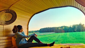 A person sitting on a bed inside a tent-like structure with a view of a green field and trees. - Under Canvas Bornholm (Østermarie)