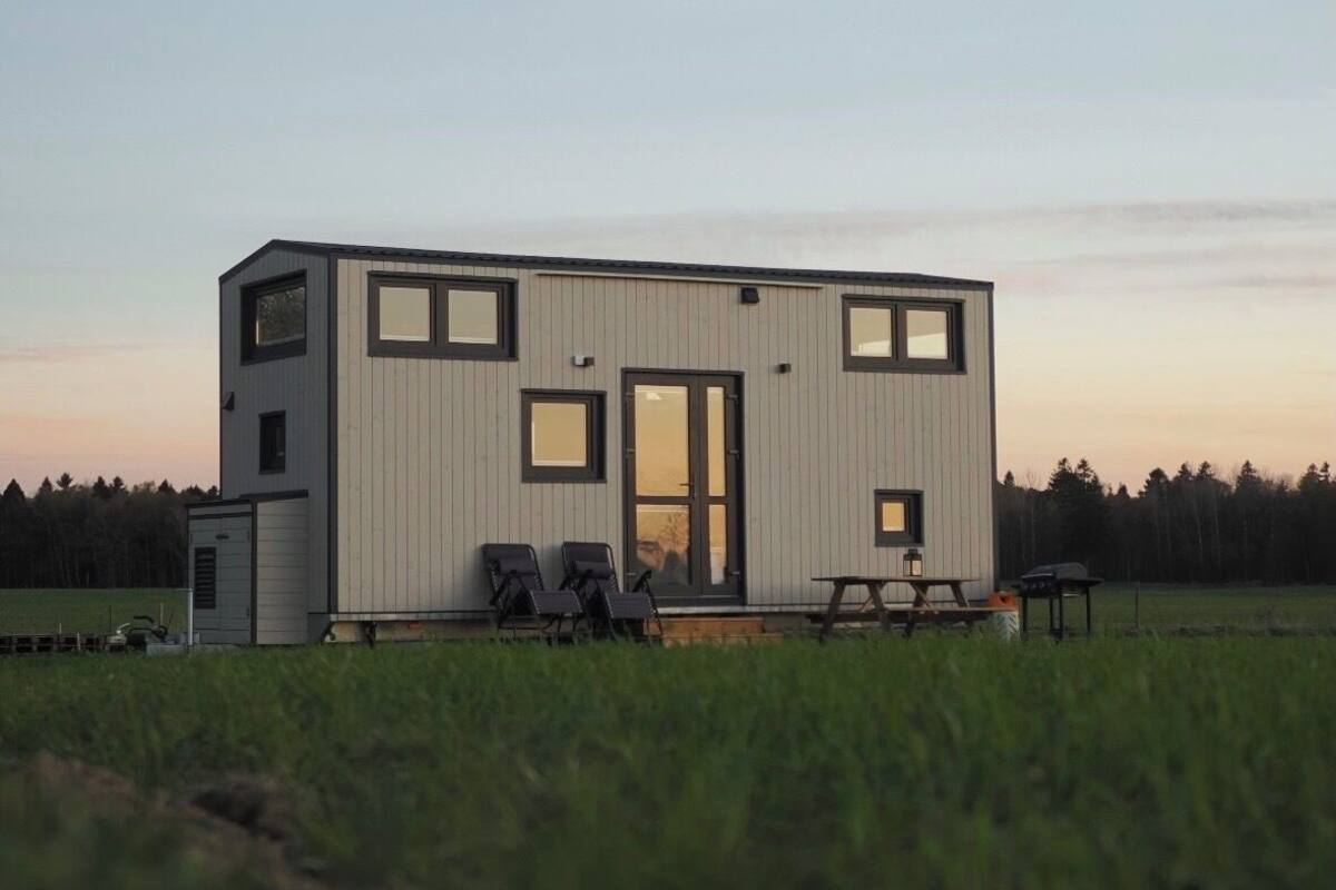 Un edificio moderno de una sola planta con techo plano, ventanas amplias y una puerta. Hay zonas de estar al aire libre con sillas y una mesa.