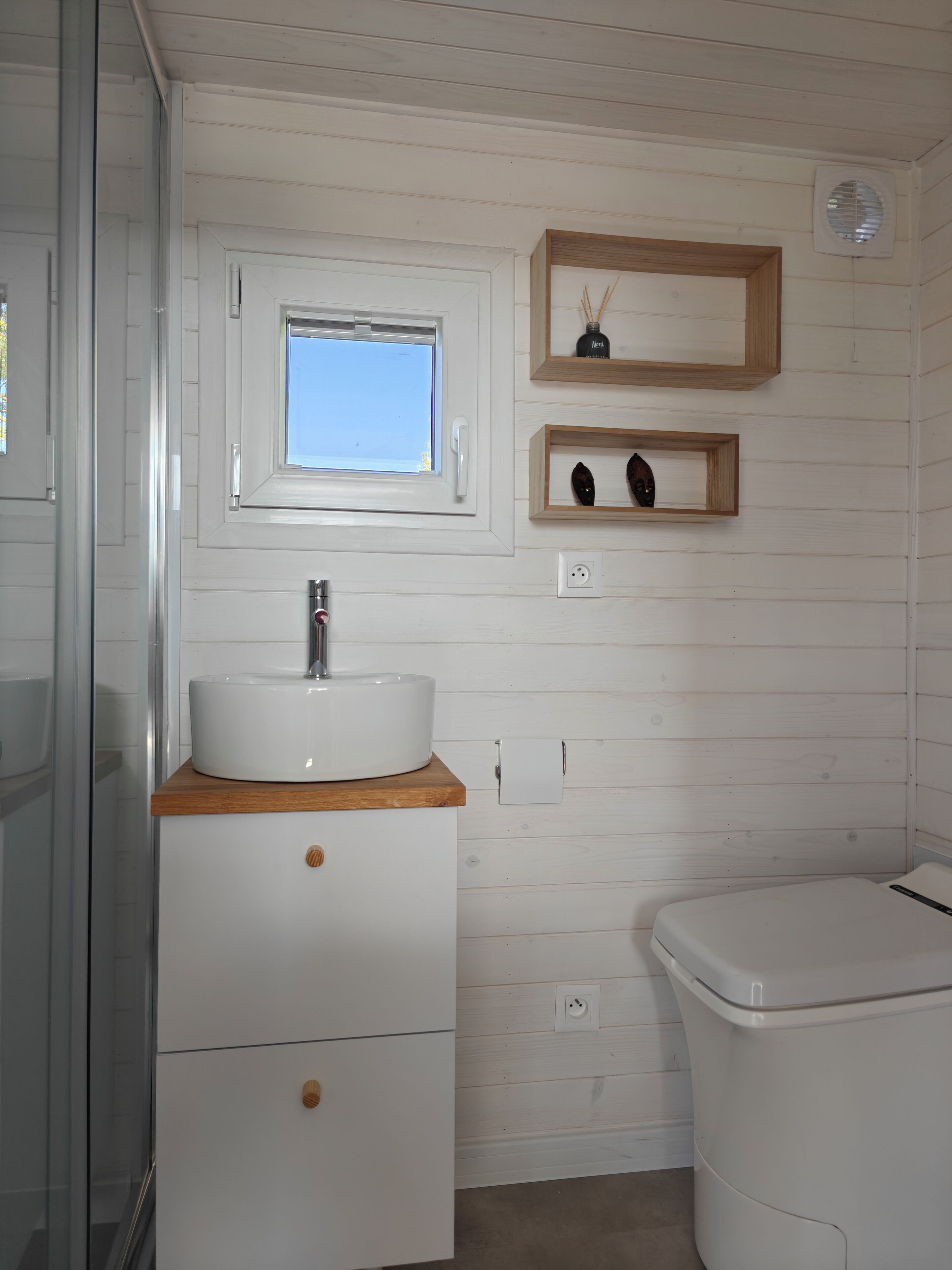 Un baño con lavamanos blanco, encimera de madera y retrete blanco. Hay una ventana sobre el lavamanos y una repisa con objetos decorativos.