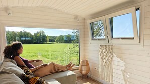 A room with a large window, a sofa, a chair, and a macrame wall hanging. - Under Canvas Bornholm (Østermarie)