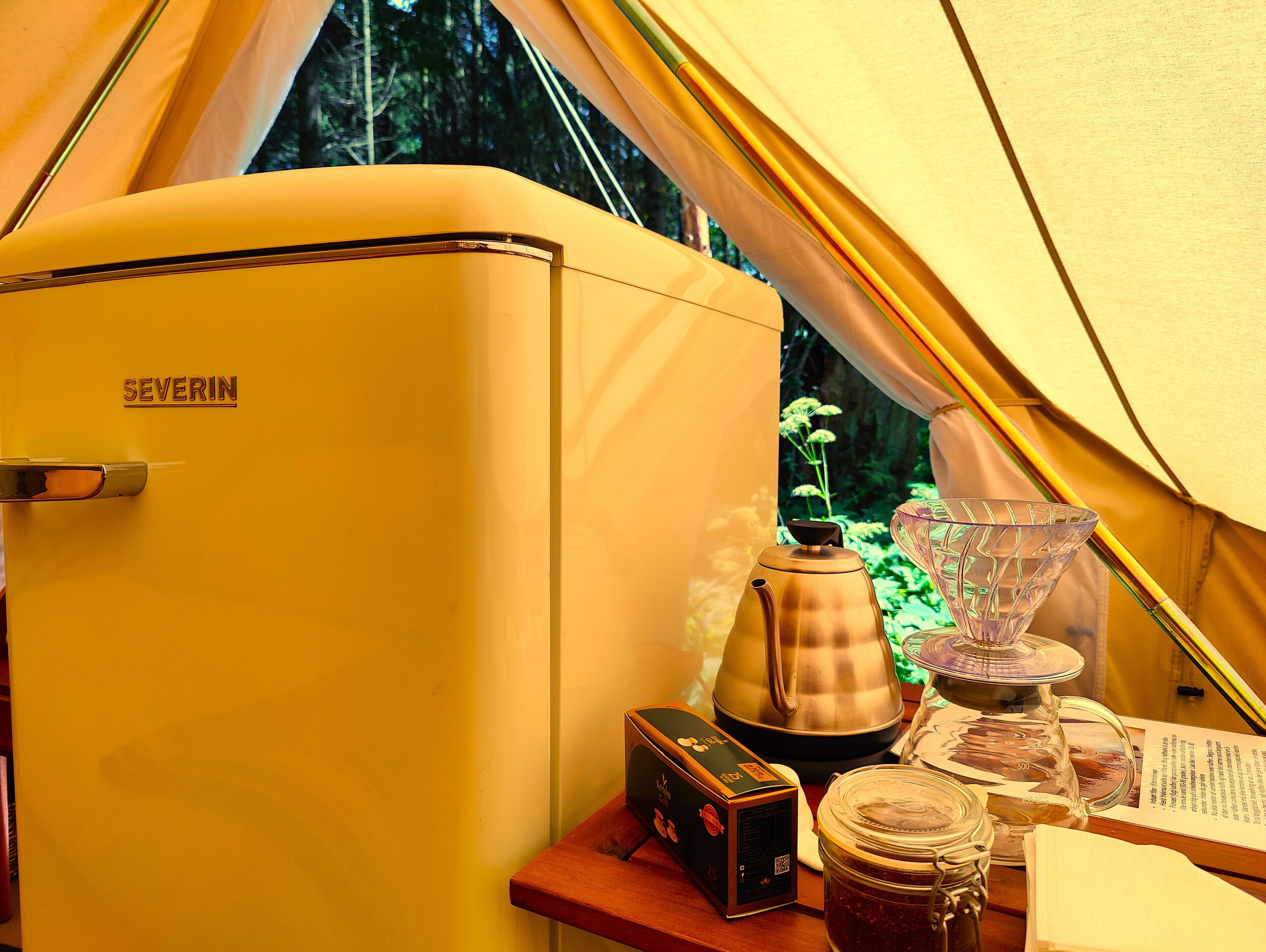 Un refrigerador amarillo antiguo con la etiqueta 'SEVERIN' dentro de una tienda de campaña, con una cafetera, un tarro de vidrio y una caja sobre la mesa.
