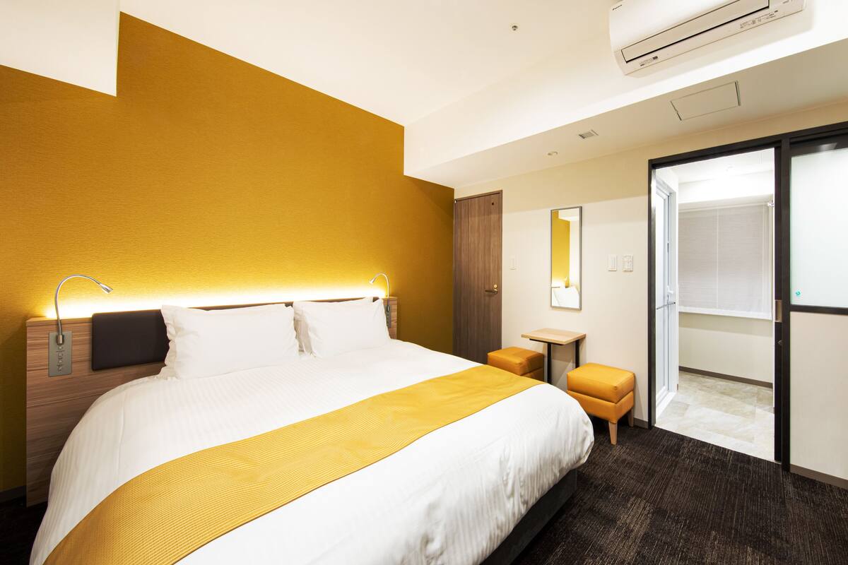 Une chambre d’hôtel avec un grand lit, une table de chevet, un appareil de climatisation, un miroir et une porte coulissante donnant sur la salle de bain.
