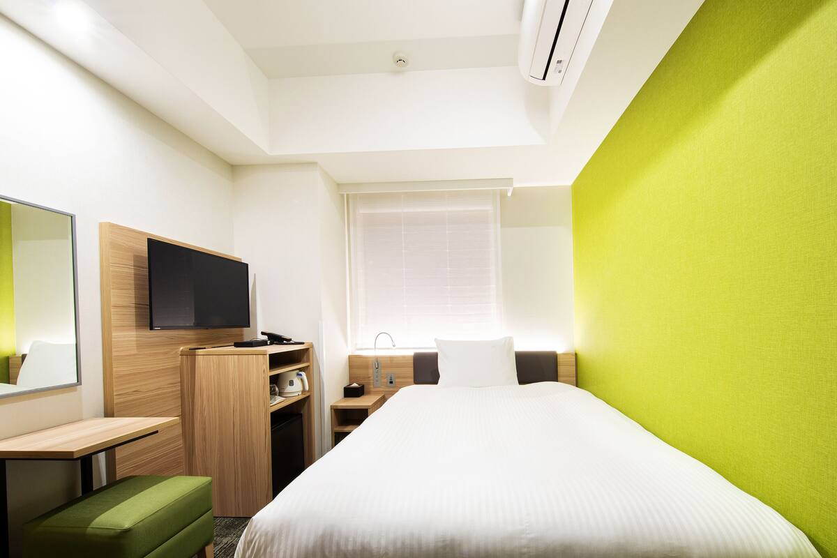 Une chambre d’hôtel avec un lit, un bureau, une télévision, un banc vert et une fenêtre avec des stores.