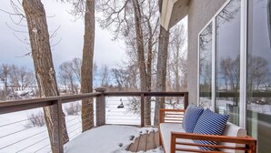 A covered deck with a wooden bench and a cushioned seat, overlooking a snowy landscape with trees and a clear sky. - Aspen Pointe Glass House on Ski Hill Road Driggs, ID (Driggs)
