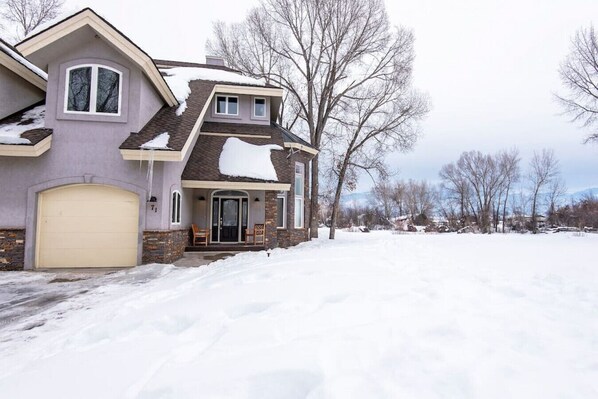 Una casa de dos pisos con un garaje, un porche delantero con sillas y un patio cubierto de nieve.