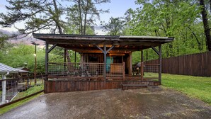 A rustic wooden structure with a covered seating area and a fenced-in space, surrounded by trees and a wooden fence. - Family Retreat, outdoor oasis, game room (Broken Bow)
