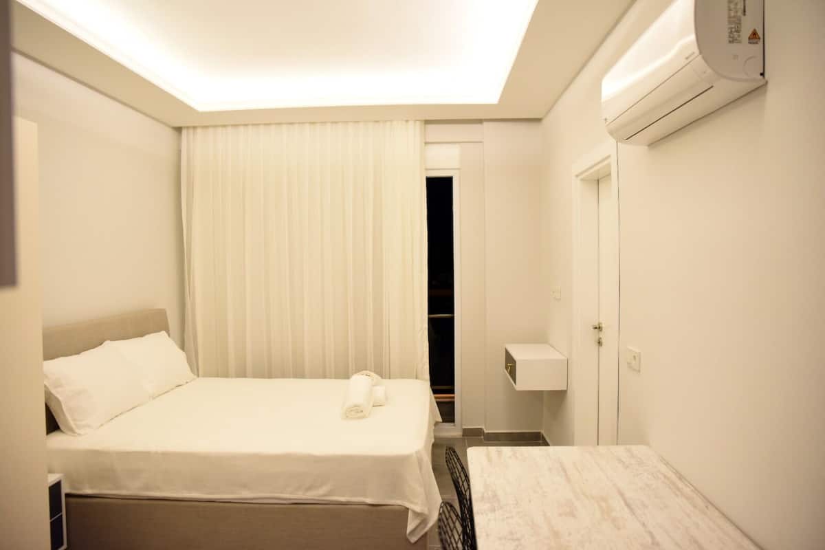 A hotel room with a bed, a bedside table, a small table, a closet, and an air conditioning unit.