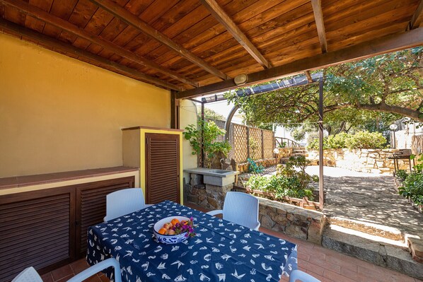 A covered patio with a table set for dining, a grill, and a view of the garden. - VILLA ALLEGRA (Villasimius)