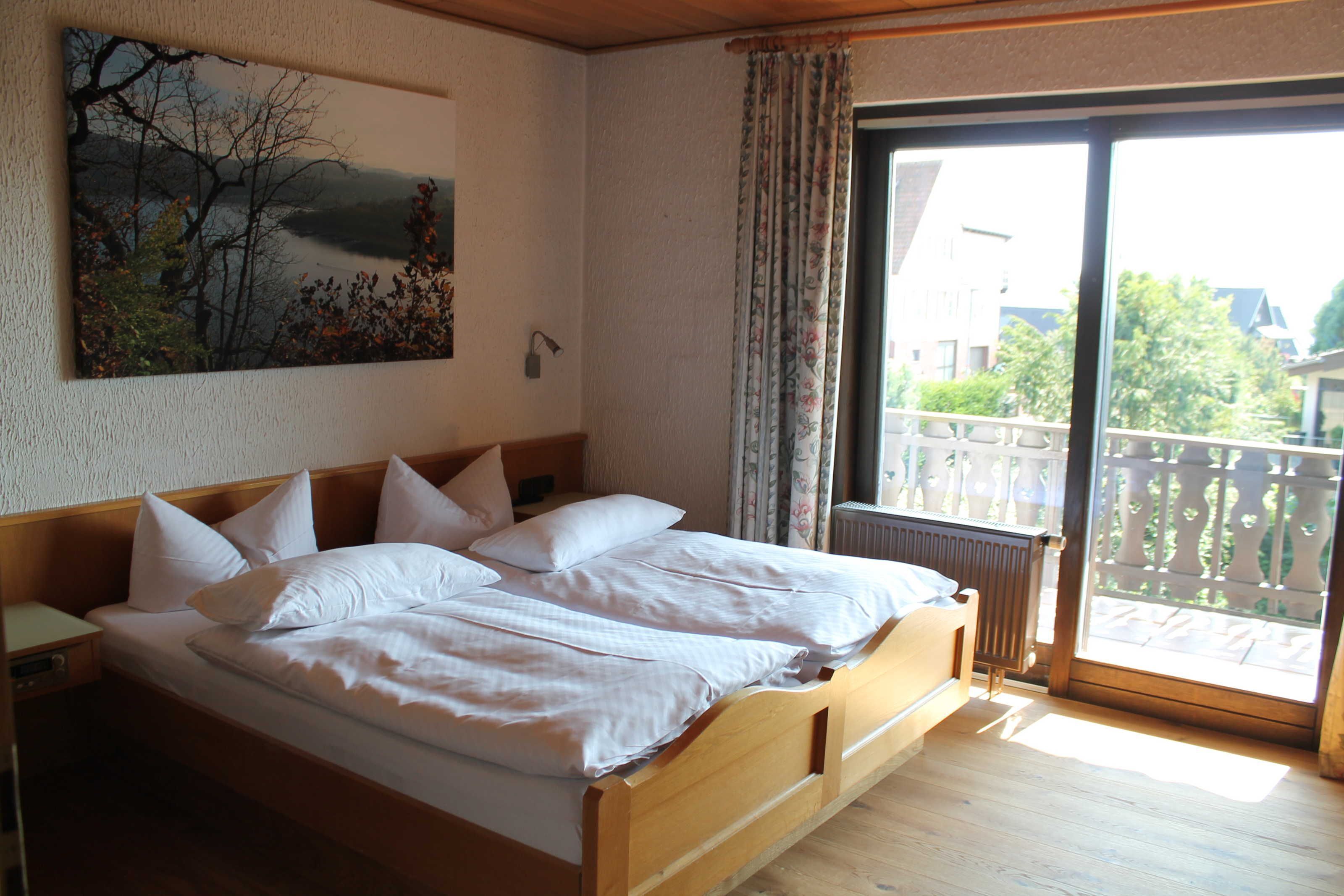 Un dormitorio con cama de madera, una mesita de noche, un cuadro en la pared y vistas a través de un ventanal grande.