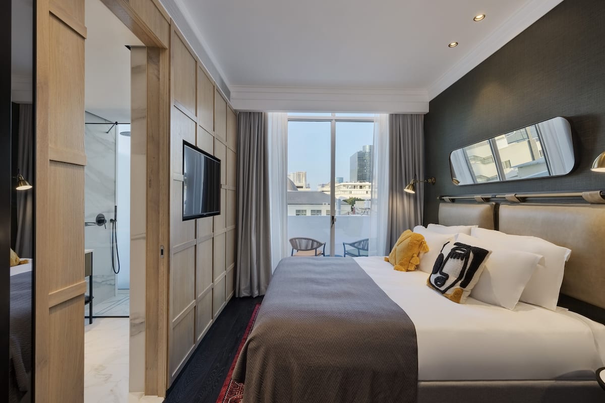 A modern hotel room with a large bed, a TV, a balcony with a view of the city, and a shower area.