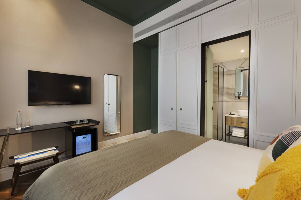 A modern hotel room with a large bed, a desk with a flat-screen TV, a mirror, and a walk-in closet.