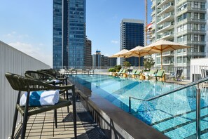 Piscina no terraço com espreguiçadeiras e guarda-sóis, cercada por edifícios altos.