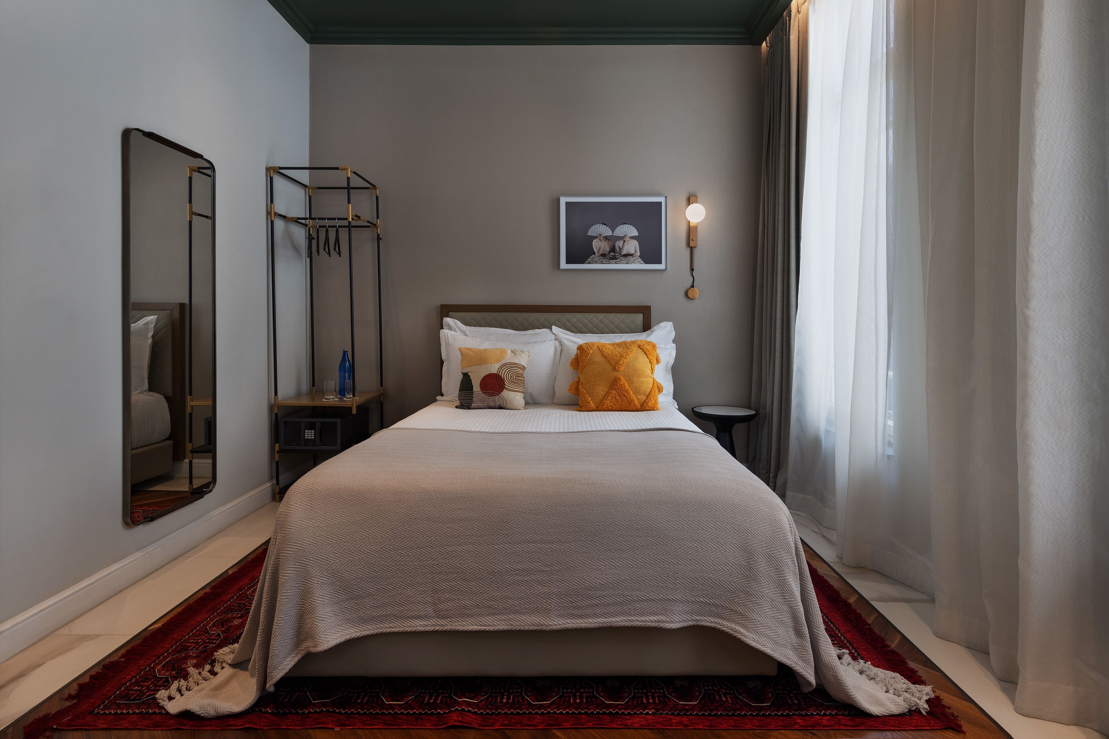 A neatly made bed with a white and beige bedspread, a red and black patterned rug, a side table with a bottle and a small lamp, a wall-mounted mirror, and a framed picture above the bed.