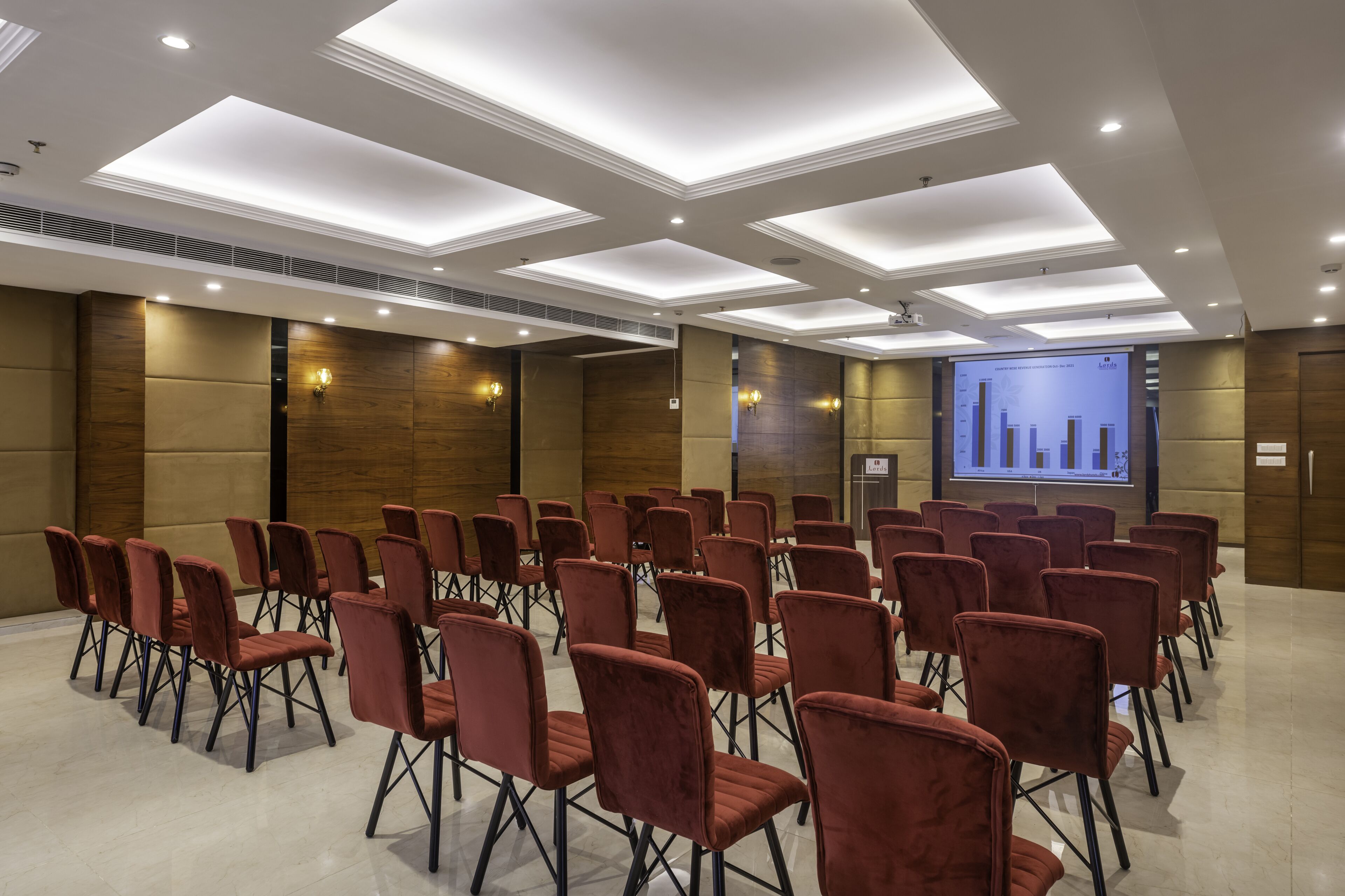 Una sala de conferències amb files de cadires vermelles, una pantalla gran que mostra una presentació i il·luminació encastrada al sostre.
