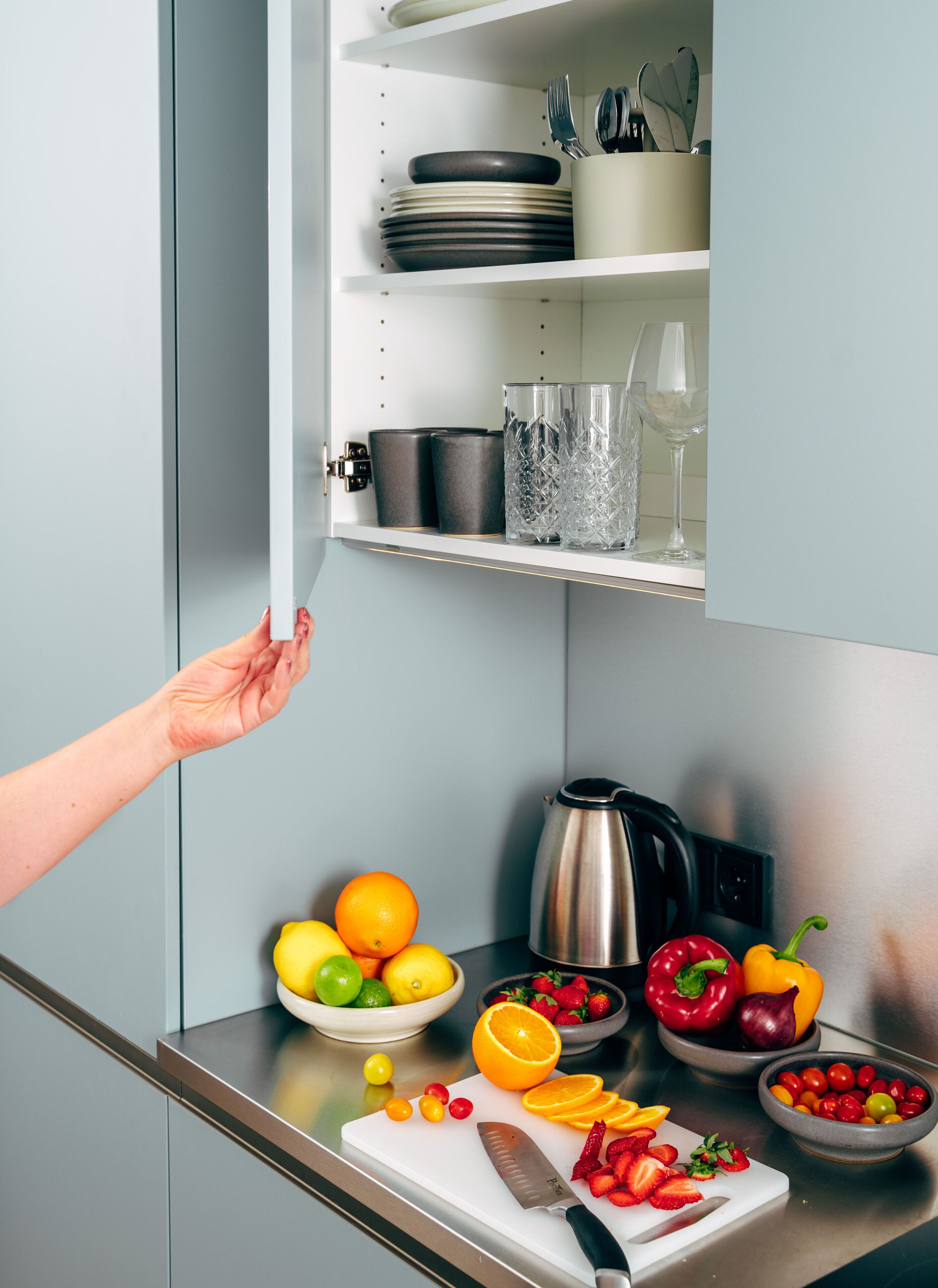 Eine Küche mit Schneidebrett, Obst, Wasserkocher und einem Schrank mit Geschirr und Gläsern.