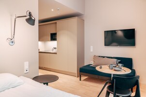 Ein modernes Hotelzimmer mit einem Bett, einem Schreibtisch, einem runden Tisch mit Stuhl, einer Couch, einem Fernseher und einer Küchenzeile im Hintergrund.