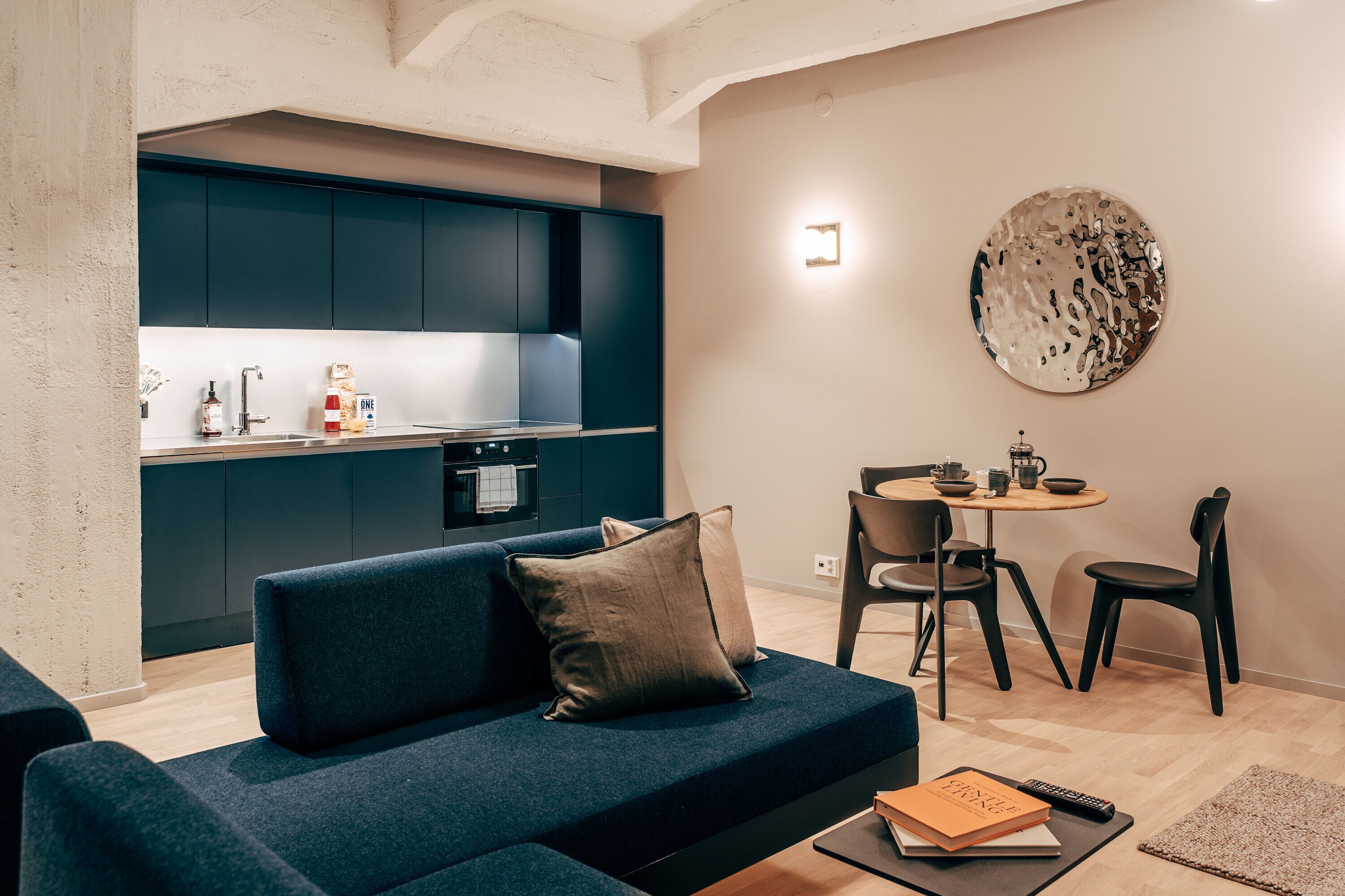 Una sala de estar moderna con un sofá azul oscuro, una mesita redonda con sillas y una zona de cocina al fondo.