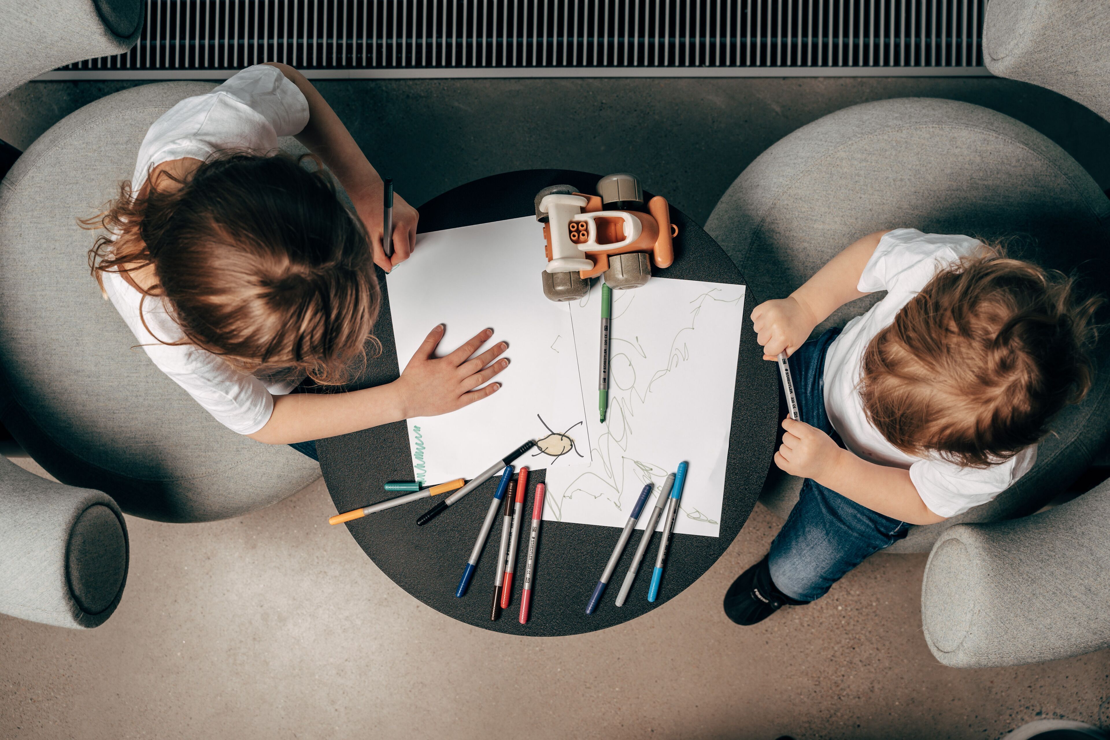 Deux enfants sont assis à une table et dessinent avec différents feutres de couleur, et une voiture jouet est posée à côté d’eux.