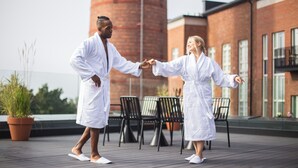 Two people in white bathrobes holding hands on a rooftop terrace with chairs and potted plants. - Unity Tampere - A Studio Hotel (Tampere)