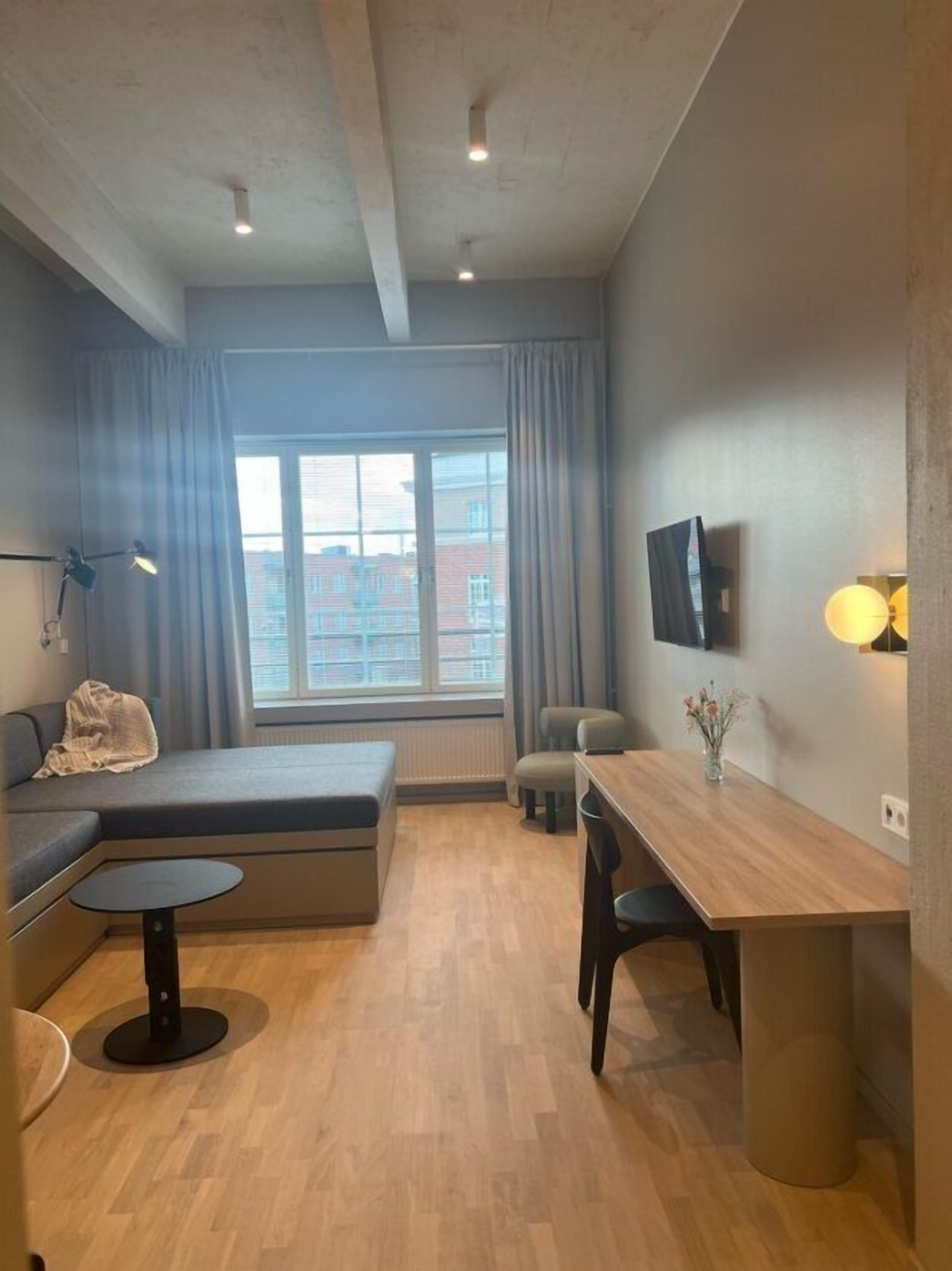 A modern, compact living space with a sofa, a small round table, a dining table, and a TV mounted on the wall.
