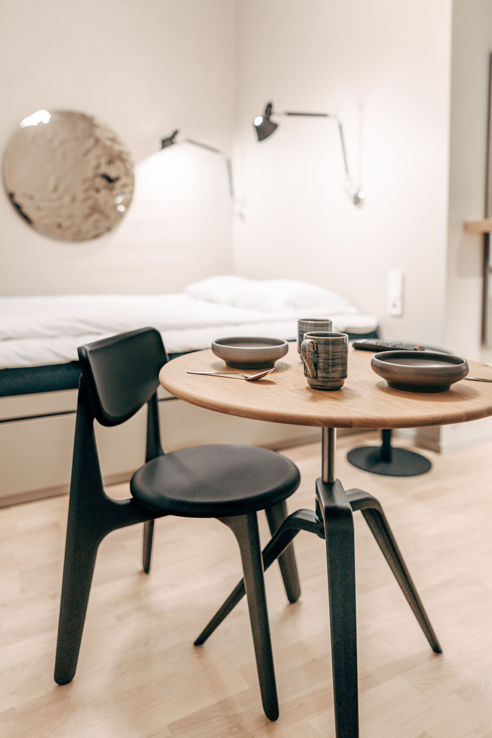 Ein modernes Zimmer mit einem runden Holztisch, schwarzen Stühlen und einem Bett im Hintergrund.
