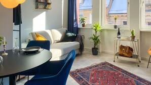 A modern living room with a sofa, a dining table, and chairs. There are potted plants, a rug, and a small table with a bottle and glass. - Astoria Apartments (Nuremberg)