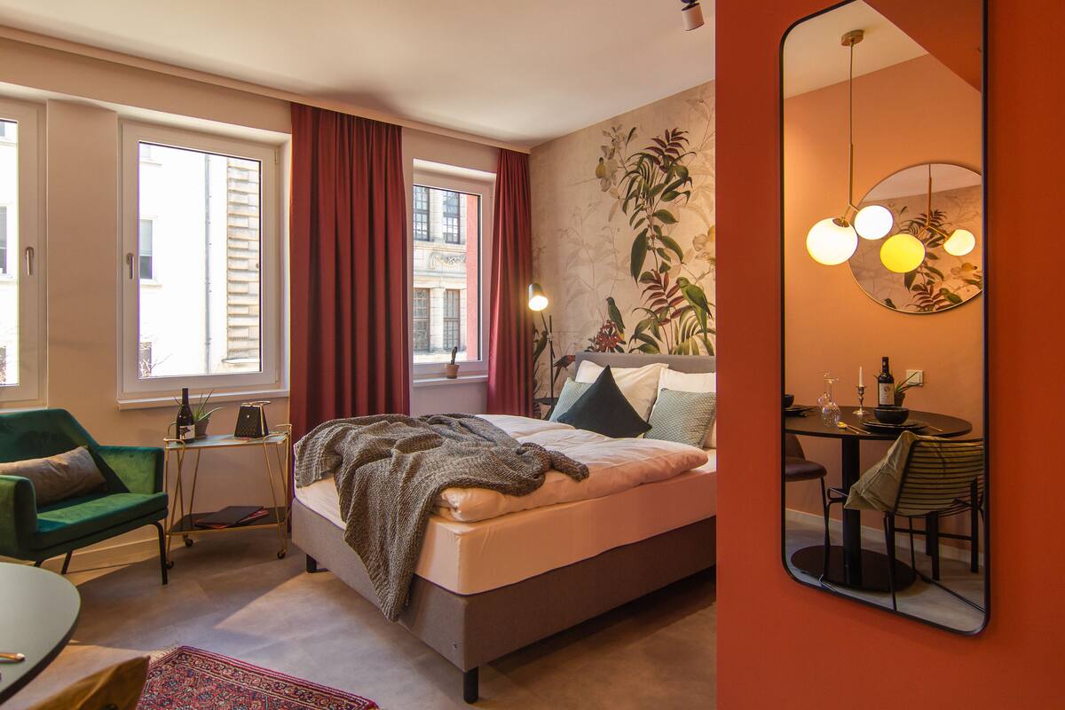 Une chambre à coucher avec un lit, un fauteuil vert, une petite table avec une bouteille et des verres, et une fenêtre avec des rideaux.