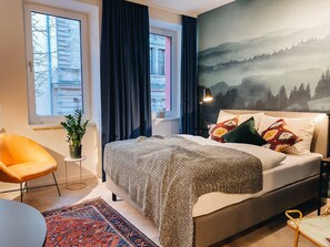 A modern bedroom with a bed, a chair, a rug, and a view of a building through the window. - Astoria Apartments (Nuremberg)